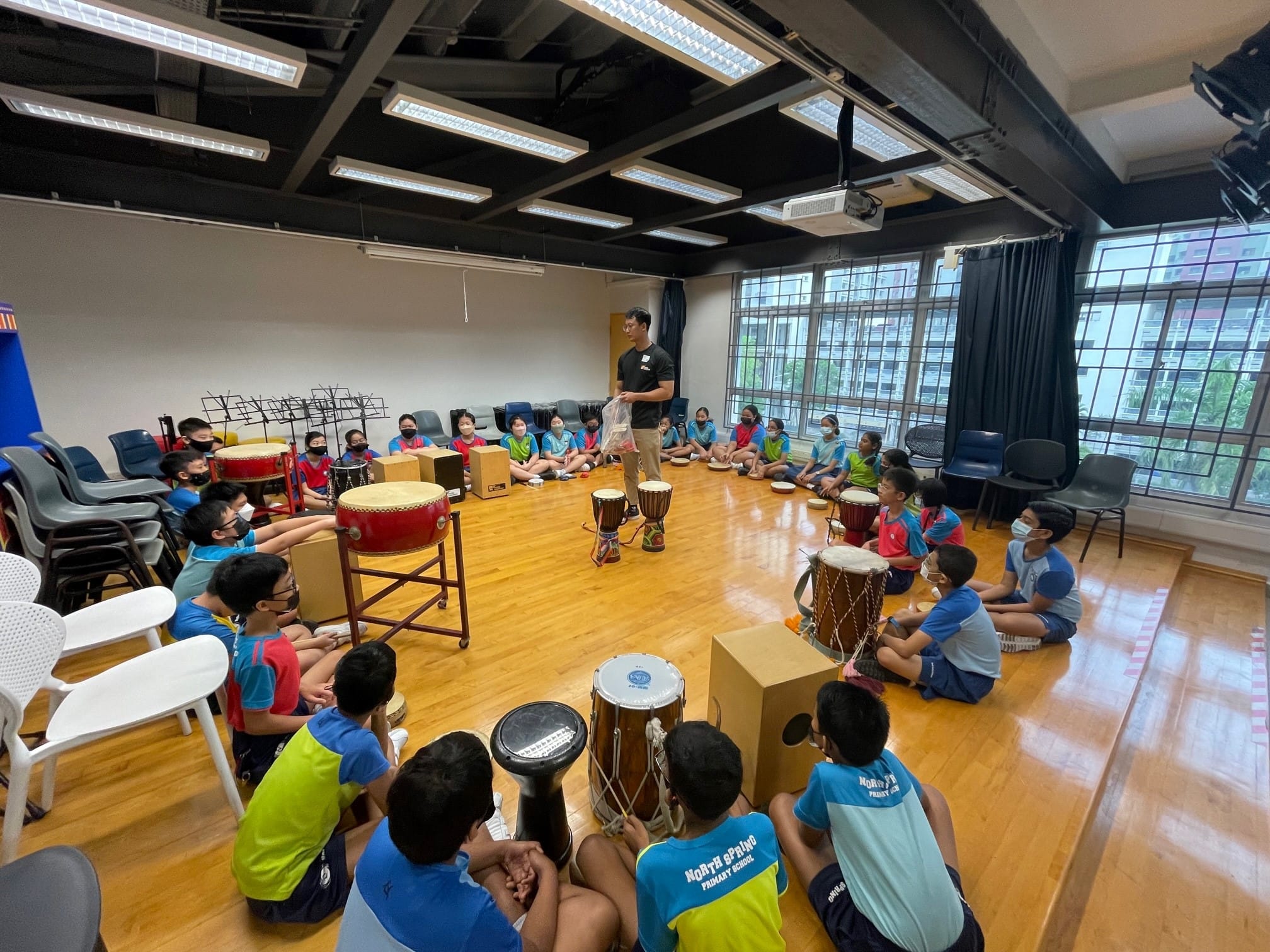 Children in school uniforms sit in a circle on a wooden floor around various drums while a teacher stands in the center.